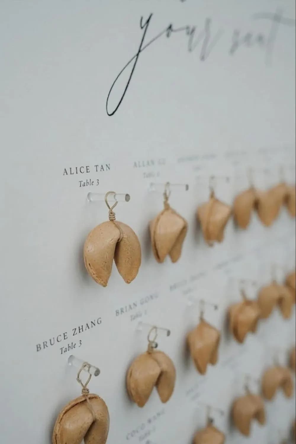 Fortune-cookie wedding seating chart: real fortune cookies hang from clear pegs on a crisp white board, each beside a guest name and table number