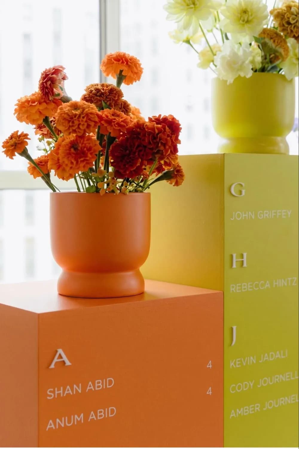 Minimalist wedding seating chart on freestanding plinth columns painted saturated orange and chartreuse, guest names laid out alphabetically, topped with marigolds in coordinating vases