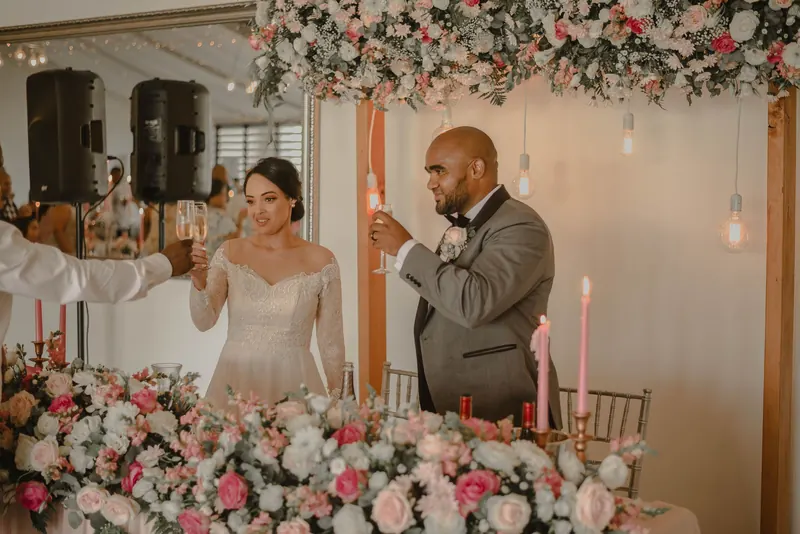 Bride and groom toasting behind a sweetheart table covered in pink and white roses with a flower wall behind them