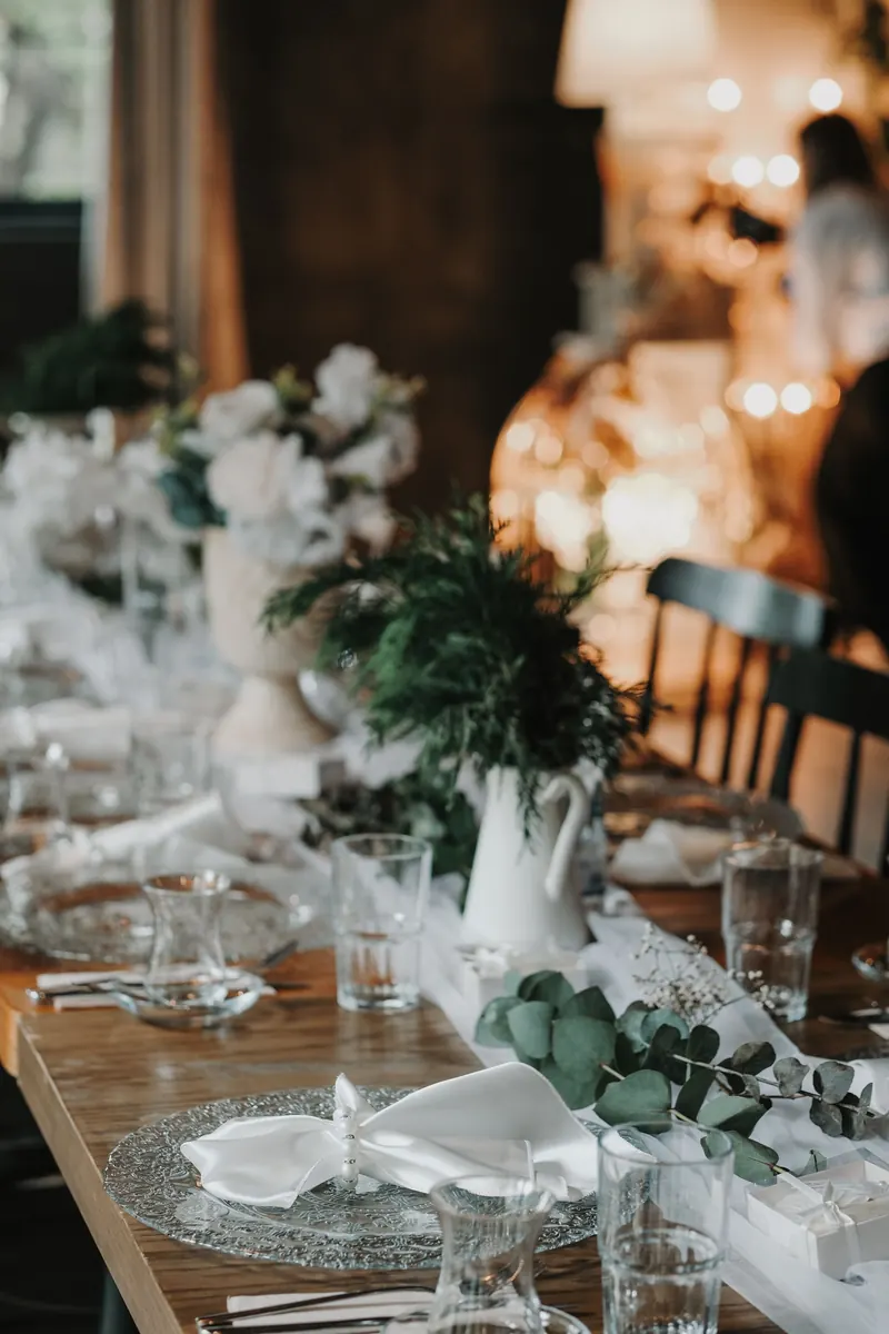 Long wedding table with a low runner of white flowers and greenery in white jugs between glassware and napkins