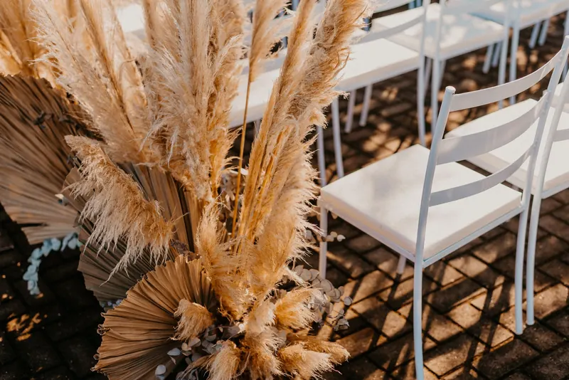 Large arrangement of pampas grass and dried palm leaves beside white chairs at an outdoor wedding