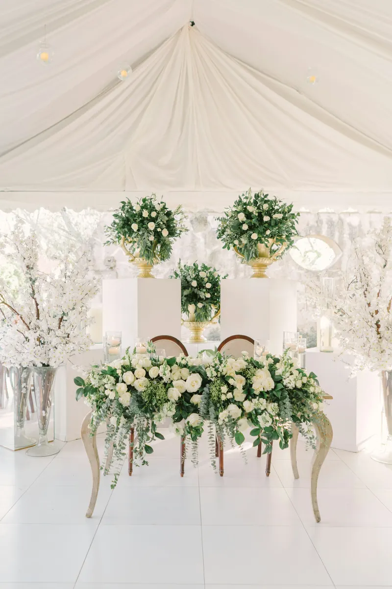 Sweetheart table for two under a white tent with white flower arrangements on the table and greenery hanging above