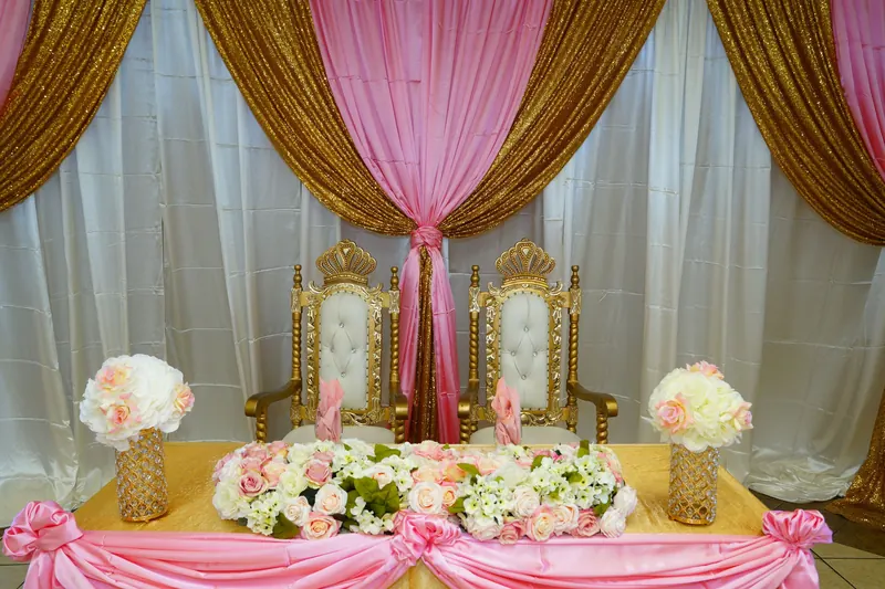 Sweetheart table with gold throne chairs, pink and white flowers and pink, gold and white fabric drapes behind