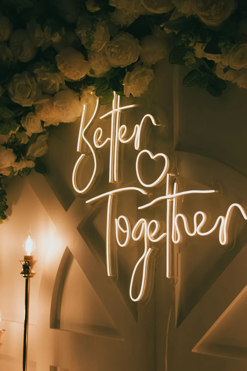 Warm white neon sign reading Better Together beneath an arrangement of white roses and greenery