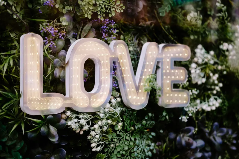 Light-up marquee letters spelling LOVE hanging on a wall of greenery and flowers