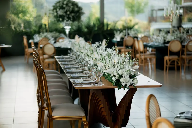 Wedding reception table lined with white flowers in a glass-walled room overlooking a garden