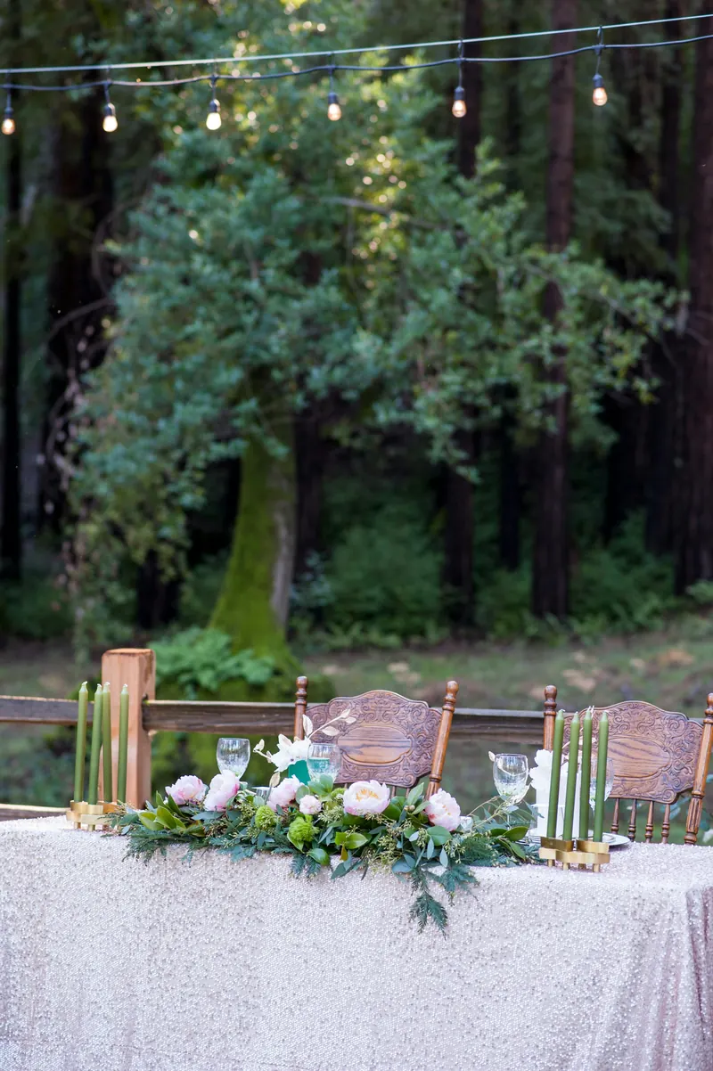 Outdoor sweetheart table in a forest with a white cloth, a greenery runner and tall green taper candles