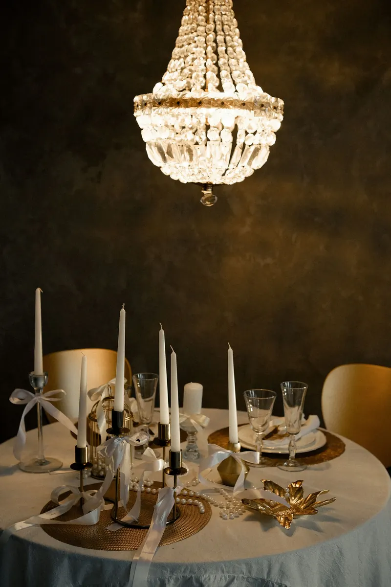 Crystal chandelier hanging over a round table set with white taper candles, ribbons and glassware