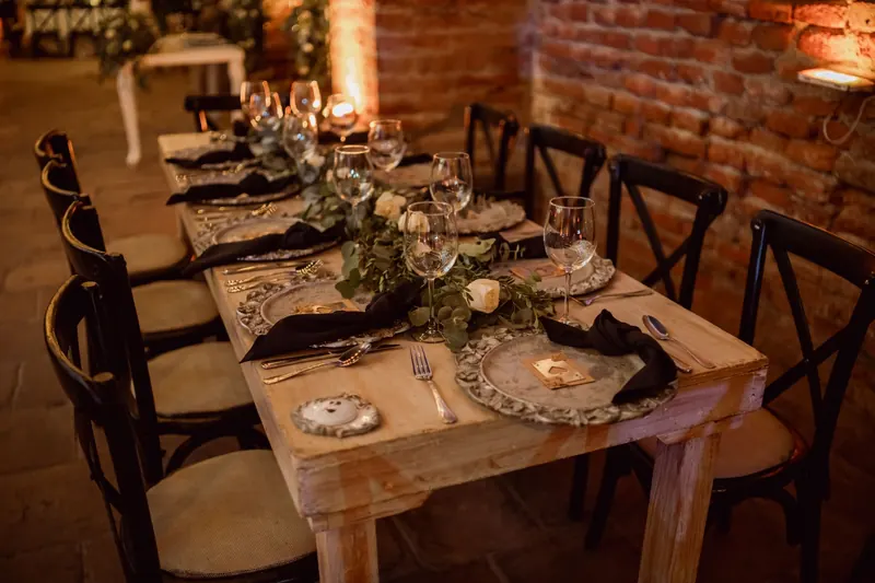 Rustic wooden wedding table with black napkins, greenery and cross-back chairs against a brick wall