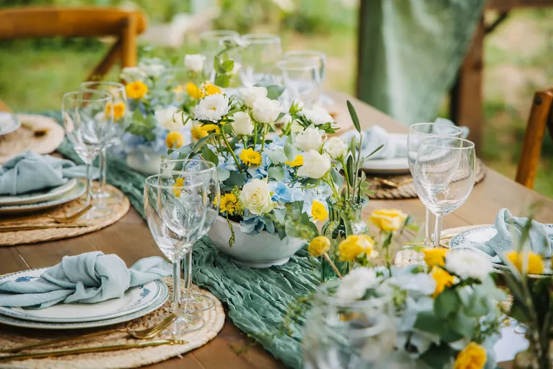Outdoor wedding table with yellow and white flowers, greenery, blue napkins and woven chargers