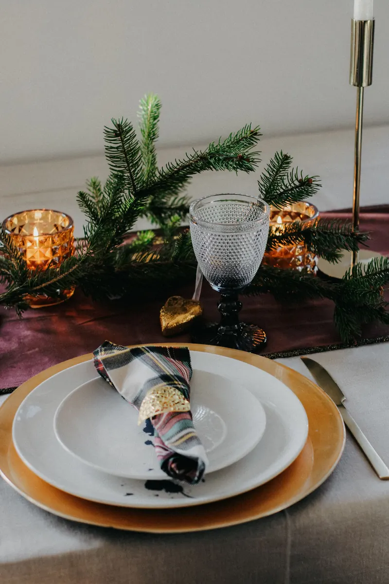 Winter table setting with a gold charger, white plates, pine branches and a candle in a glass holder