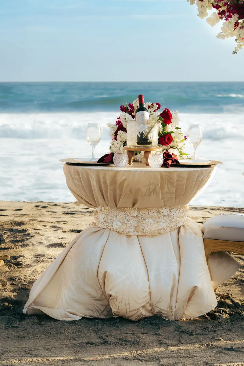 Small round table draped in white linen with a flower arrangement on a sandy beach with the ocean behind
