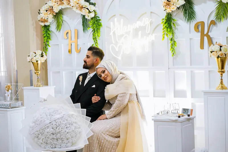 Bride and groom seated in front of a white backdrop with floral sprays and large gold initial letters on stands
