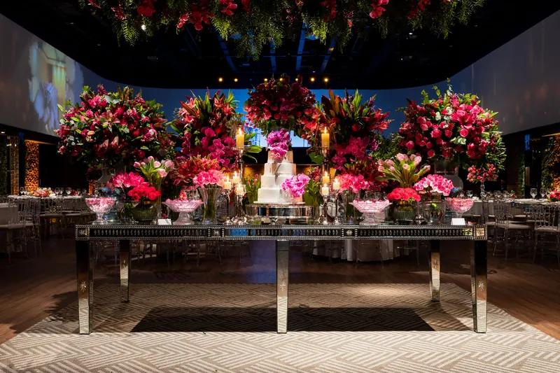 Wedding cake on a mirrored table surrounded by vibrant pink and red flower arrangements